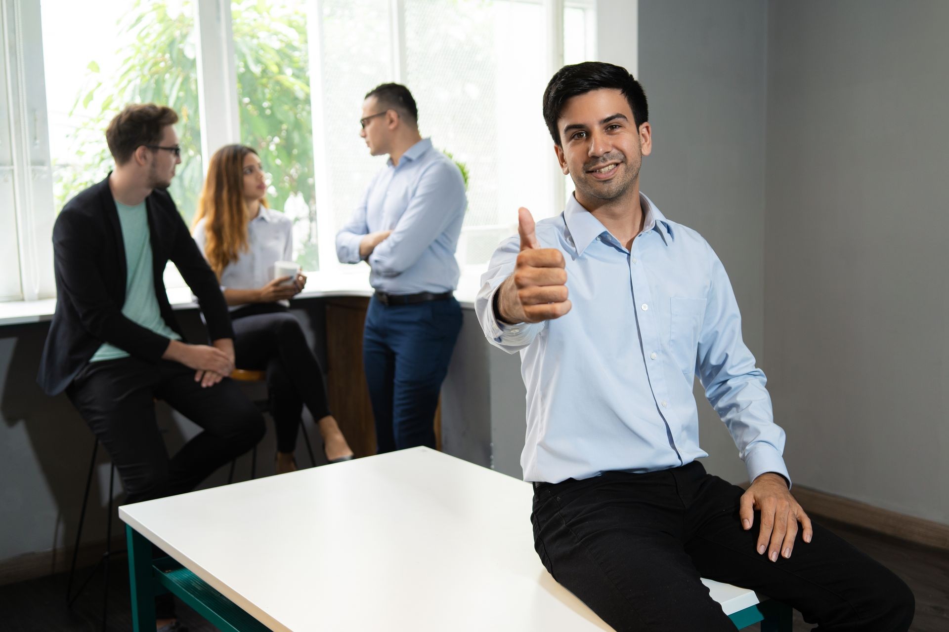Positive manager proud of his job and team. Smiling office employee giving thumb up at camera, coworkers drinking coffee and chatting in background. Approval and good job concept