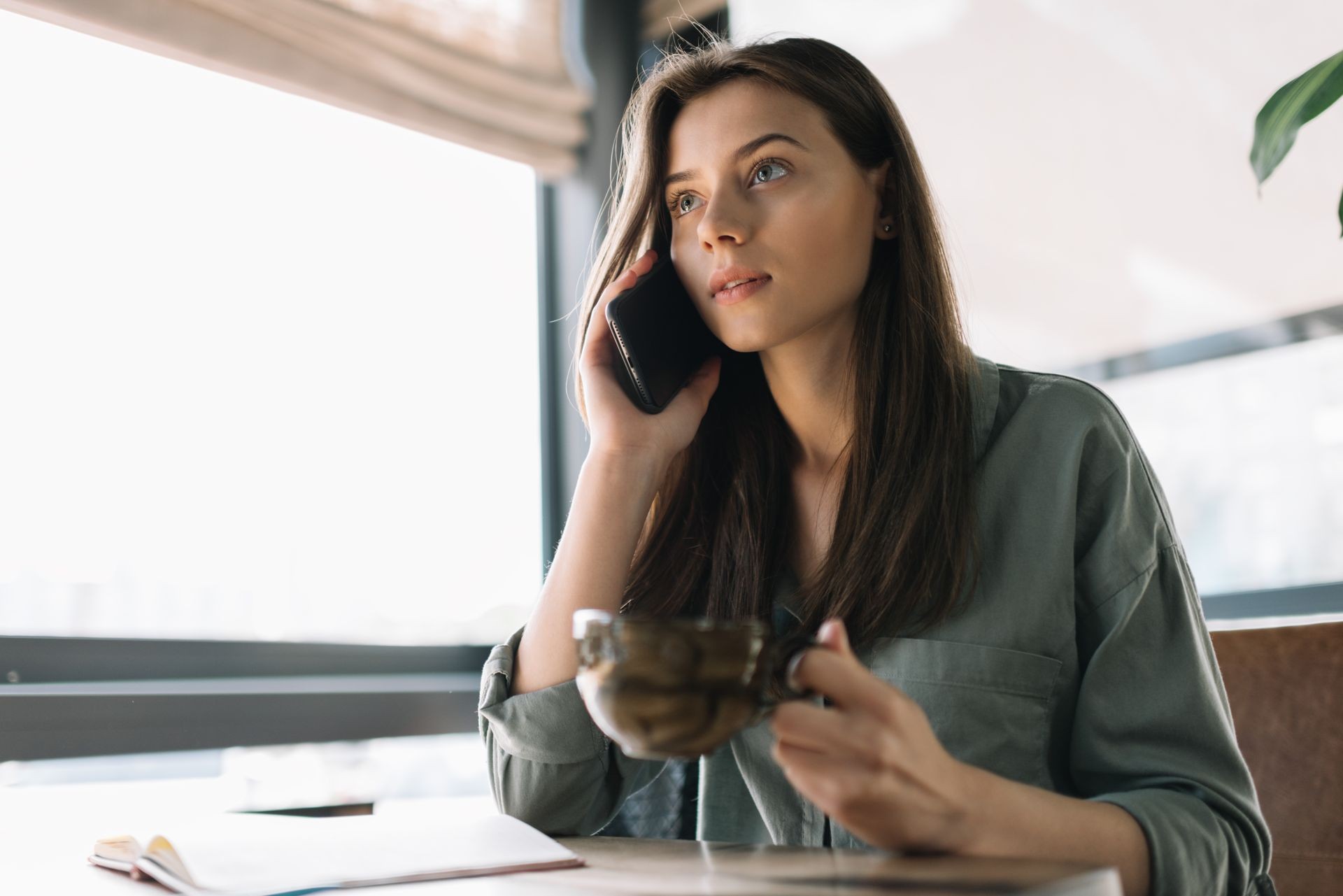 Attractive caucasian businesswoman holding smartphone and cup of coffee sitting at office. Young beautiful female using mobile phone in modern coffee shop.